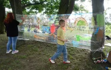 dziecko i nauczyciel malują farbami na folii rozpiętej pomiędzy drzewami