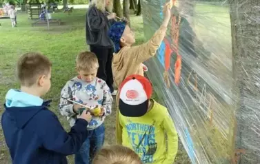 dzieci malują farbami na folii rozpiętej pomiędzy drzewami