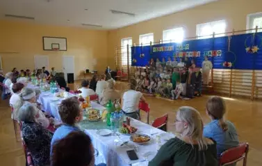 Seniorzy z Dziennego Domu Pobytu przy stole, w tle dzieci przedstawiający program z okazji Dnia Rodziny