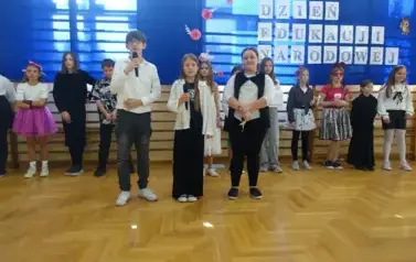 przedstawienie uczni&oacute;w klas IV i V z okazji Dnia Edukacji Narodowej, na pierwszym planie troje uczni&oacute;w składających życzenia