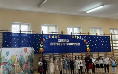 dzieci z oddziału przedszkolnego stojące w p&oacute;łkolu na tle napisu Dzień Babci i Dziadka