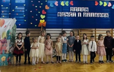dzieci z oddziału przedszkolnego stojące w p&oacute;łkolu na tle napisu Dzień Babci i Dziadka