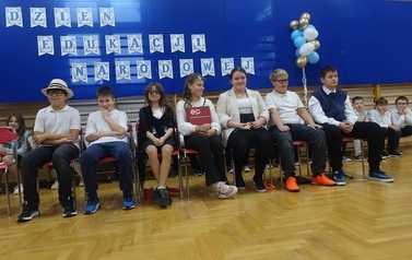 Uczniowie przedstawiają scenkę siedząc na krzesłach podczas szkolnych obchod&oacute;w Dnia Edukacji Narodowej.