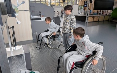 Dw&oacute;jka chłopc&oacute;w korzystających ze stacjonarnych w&oacute;zk&oacute;w inwalidzkich, trzeci chłopiec przygląda się im