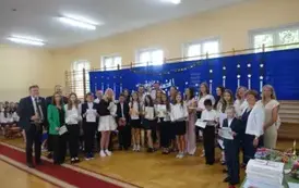 uczniowie trzymający świadectwa z wyr&oacute;żnieniem wraz z rodzicami, wychowawcą, Panią Dyrektor i Panem Burmistrzem