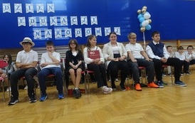 Uczniowie przedstawiają scenkę siedząc na krzesłach podczas szkolnych obchod&oacute;w Dnia Edukacji Narodowej.